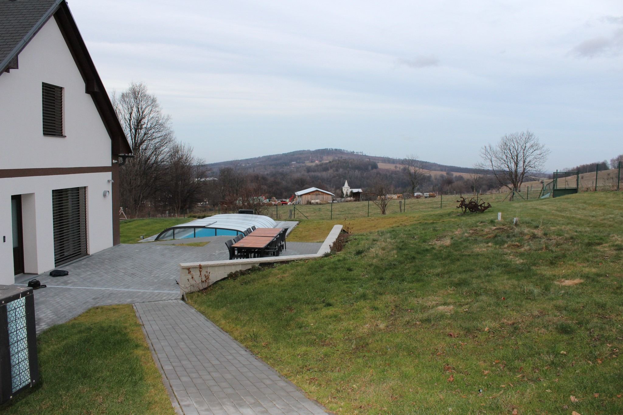 Roh domu, dlážděná cesta k zakrytému bazénu a krajina v předjaří